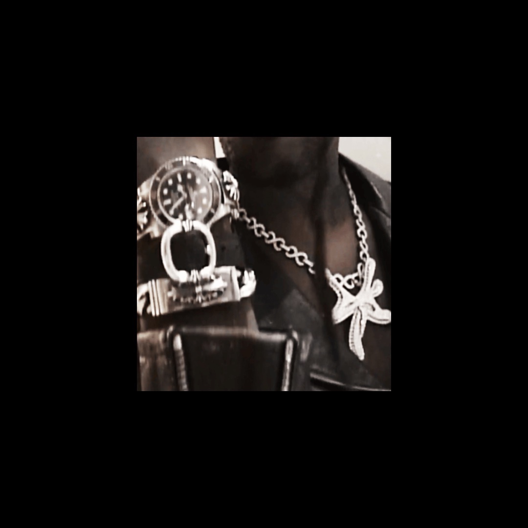 Close-up of a person’s accessories, including a large silver-toned wristwatch with a dark face, a chunky chain bracelet and a necklace with a star-shaped pendant.