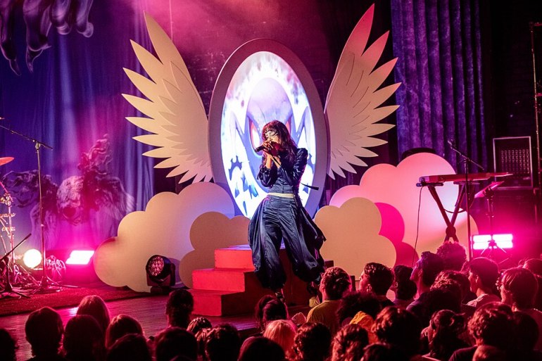 Woman singing into microphone on stage in front of lights with wings and clouds.