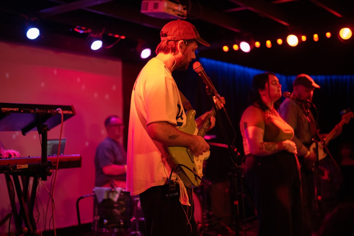 Side view of Tokyo Tea Room performing on the stage. Guitarit in the middle, lead singer on right, and drummer in the back