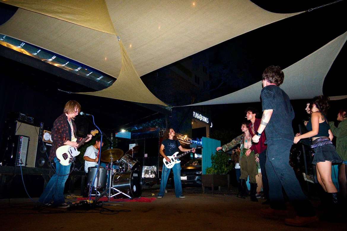The image shows a wide show of the three members of Lilium playing onstage on the left while five people from the crowd are shown on the right side dancing. Various triangle shaped tarps cover them overhead and the background shows a few amps and a building in the back.