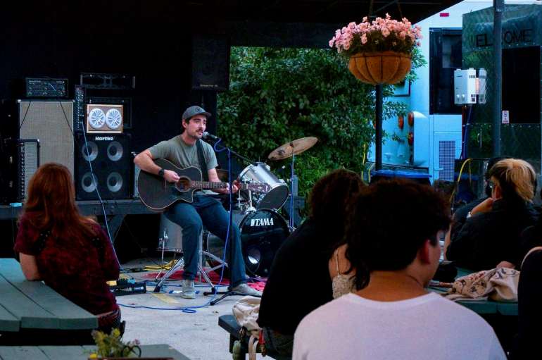 The image shows a wide shot of Dylan Hunter sitting on a stool playing a black acoustic guitar and singing into a microphone that has a blue cord around it and various amps and a drum kit are shown in the background. Some members of the crowd are sitting on gray benches in the foreground and a basket of pink flowers hangs in the top right of the image.