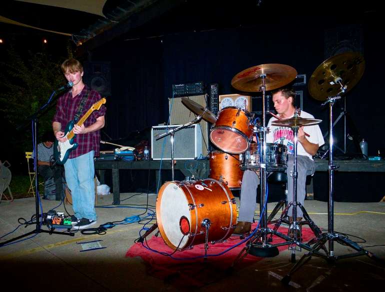 The image shows a medium ¾ shot of both members of Backswimmer, with the guitarist singing into a microphone on the left and the drummer playing on the right while a white flash shines on them and the background shows various amps.