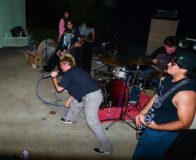 The image shows a medium ¾ shot of the four members of Cuerno performing onstage, with the guitarist on the right, vocalist in the middle, bassist on the left, and drummer in the back, while a bright white flash lights them up and two attendees stand in the background.