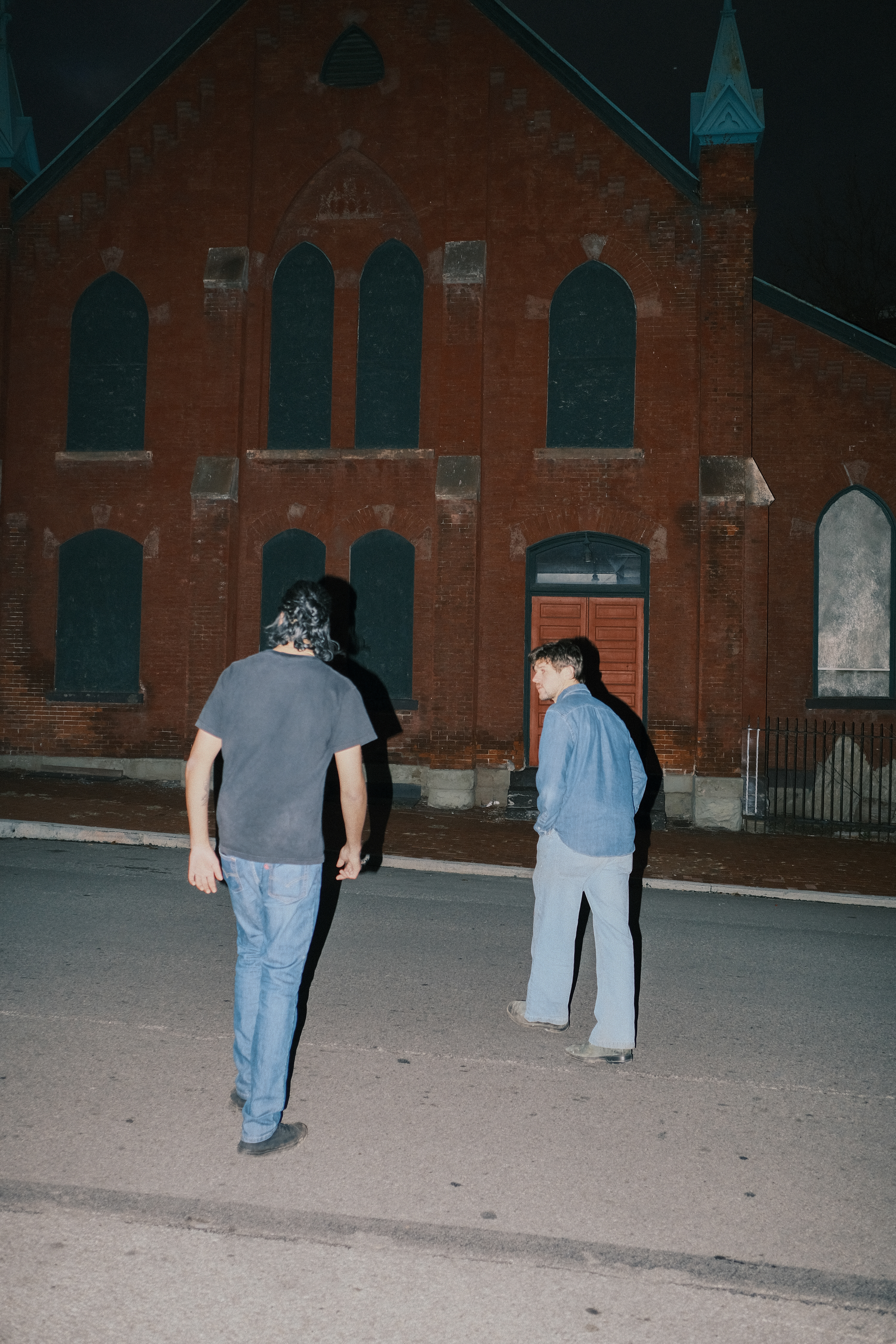 High-angle photograph of two Warren Commission band members positioned apart on a dark floor. One wears a pale pink shirt and gray trousers, while the other stands farther back in an oversized black suit with shoulder-length dark hair.