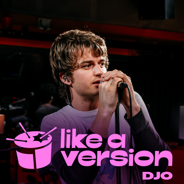 Cover for Djo’s 2025 single, Gasoline. The image is of lead singer, Joe Keery, in a grey t-shirt over a back long sleeve. Keery is holding and singing into a microphone, looking off into the right side. Keery appears to be in a recording studio with dark lighting around him. At the bottom of the cover is a purple drum and ‘like a version DJO’ in a light purple.