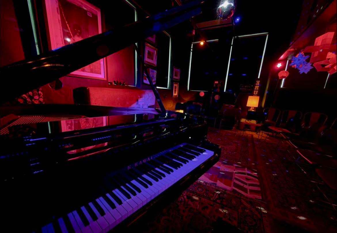 The image shows a wide shot of the inside of Blackbox, which includes low, red lighting, chairs and lamps hanging on the right side, patterned carpet on the ground, different framed photos on the left wall, a disco ball hanging overhead, and a piano on the left side of the photo.